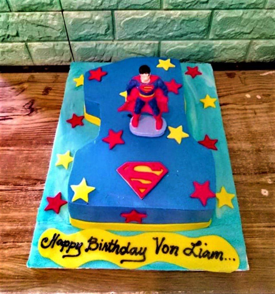 Superman themed number one shaped cake in blue with red and yellow stars and a Superman figurine.