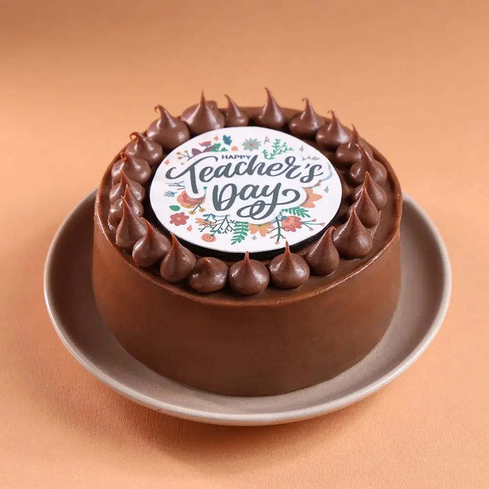 Chocolate cake with smooth brown icing, topped with a circular Happy Teacher's Day printout and chocolate icing dollops.