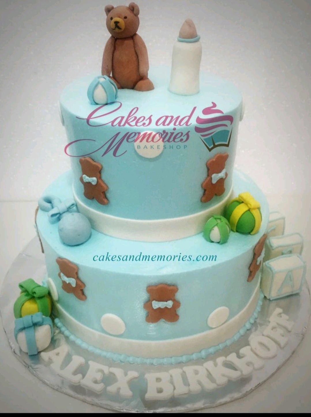 Two-tier light blue fondant baby shower cake with a brown teddy bear topper, baby bottle, and alphabet blocks.