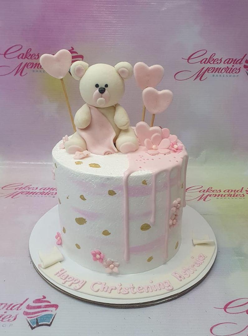 White and pink 1-tier christening cake with a large fondant teddy bear topper, pink hearts, and gold paint splatters.