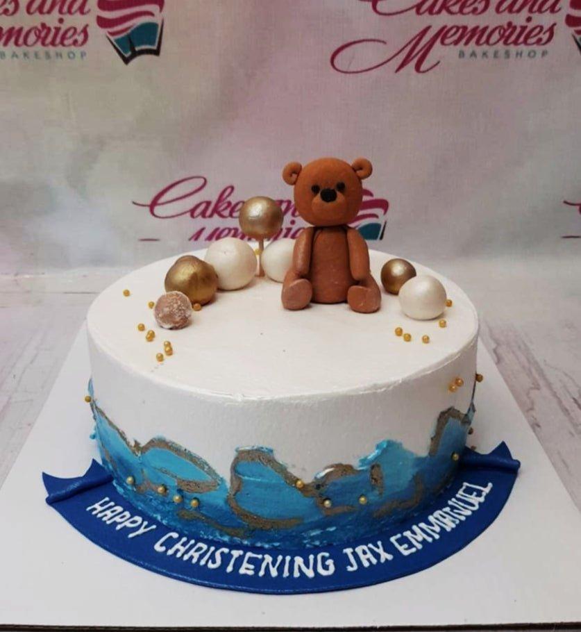 White and blue christening cake with a brown fondant teddy bear topper and gold and white decorative spheres.