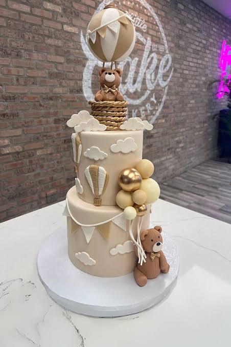 Elegant 2-tier beige fondant cake featuring a teddy bear in a hot air balloon topper, clouds, and gold balloon accents.