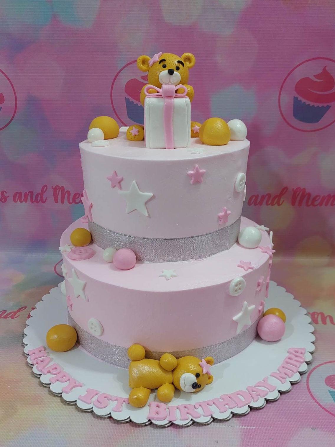 Two-tier pink birthday cake featuring a fondant teddy bear holding a gift box, decorated with stars, buttons, and silver ribbons.