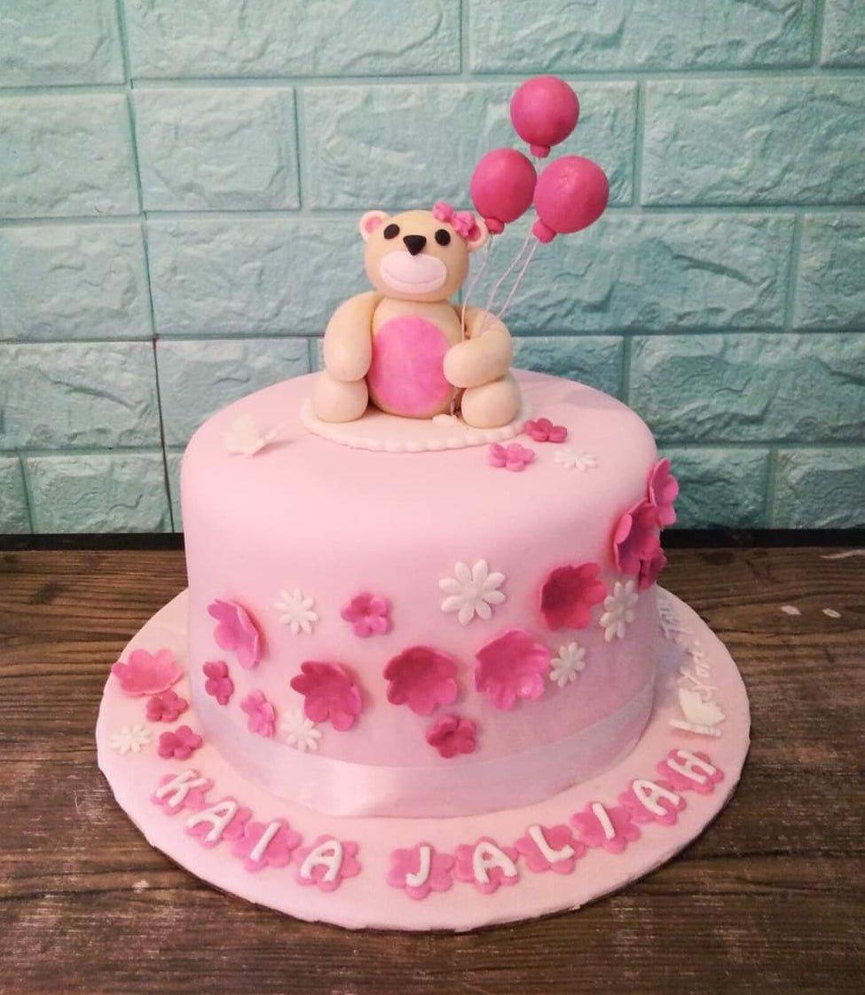 Pink fondant cake with a 3D teddy bear topper holding pink balloons and decorated with pink and white fondant flowers.