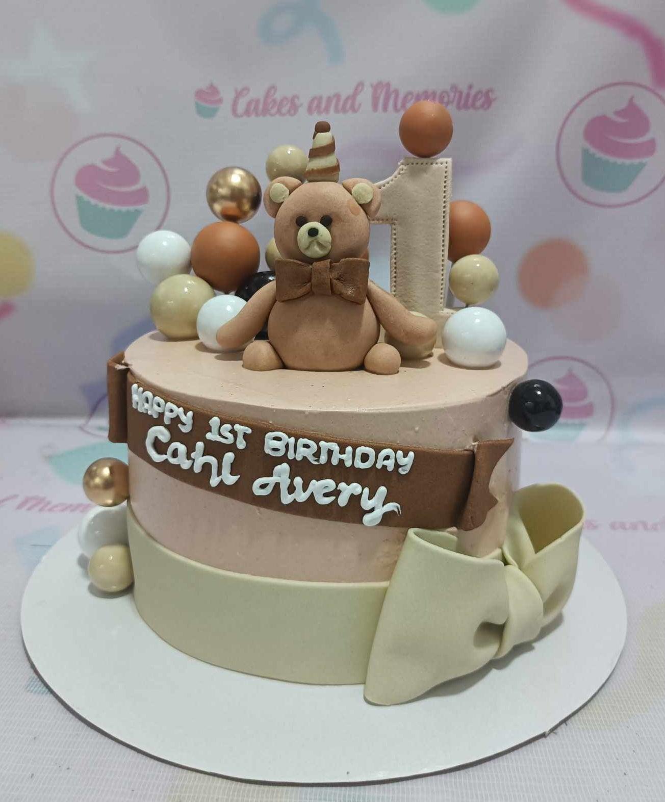 Tan 1-tier teddy bear birthday cake with a fondant bear topper, number one, and decorative spheres in brown, white, and gold.