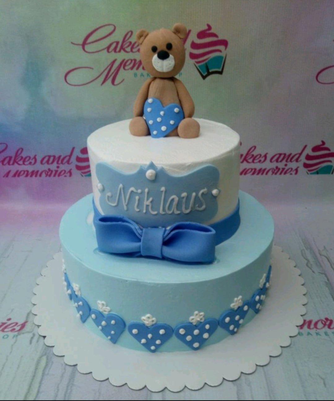 Two-tier blue and white teddy bear cake with a large blue bow and heart decorations.