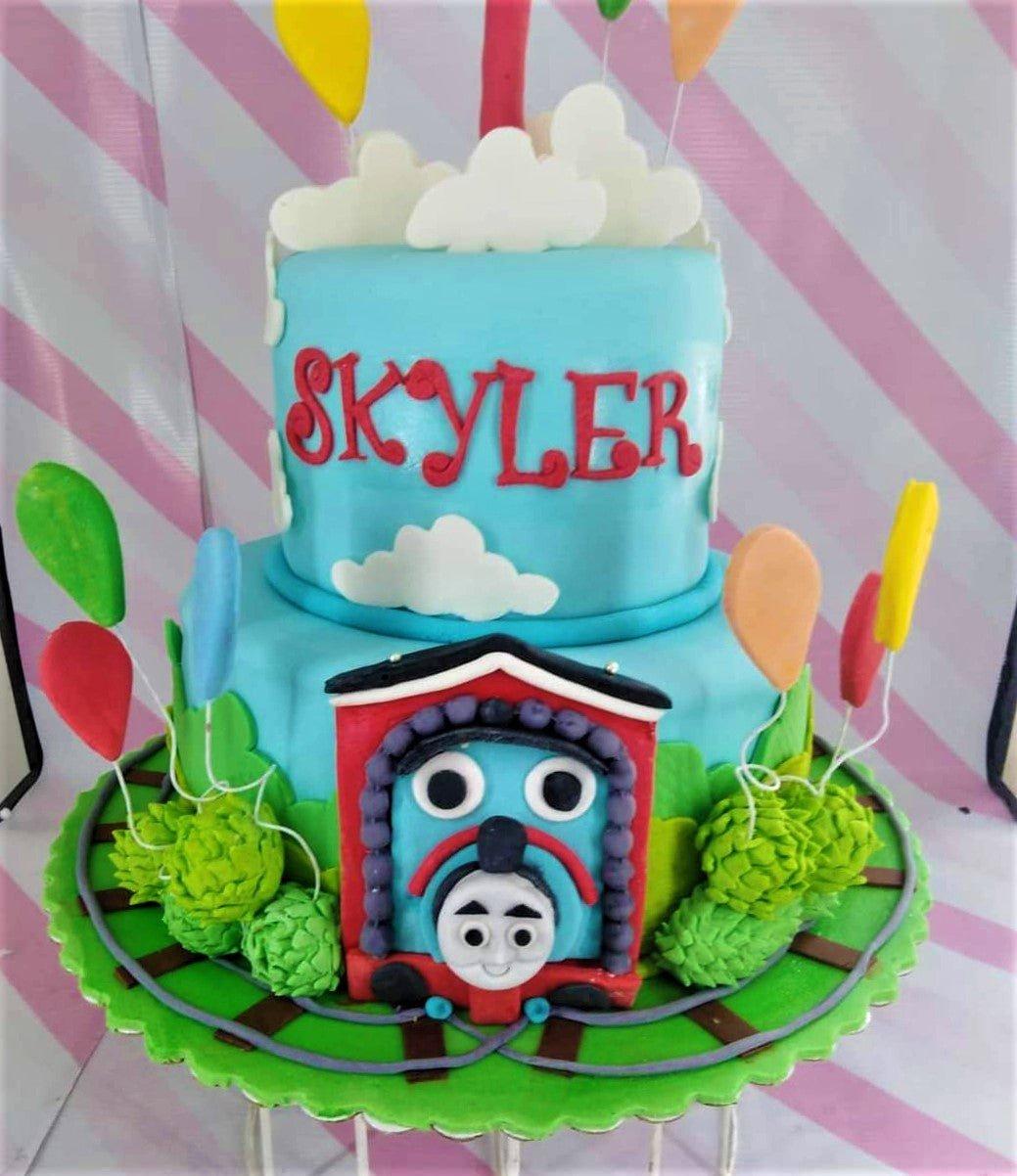 Two-tier light blue Thomas the Tank Engine themed cake with fondant clouds, balloons, and a 3D train face on the bottom tier.