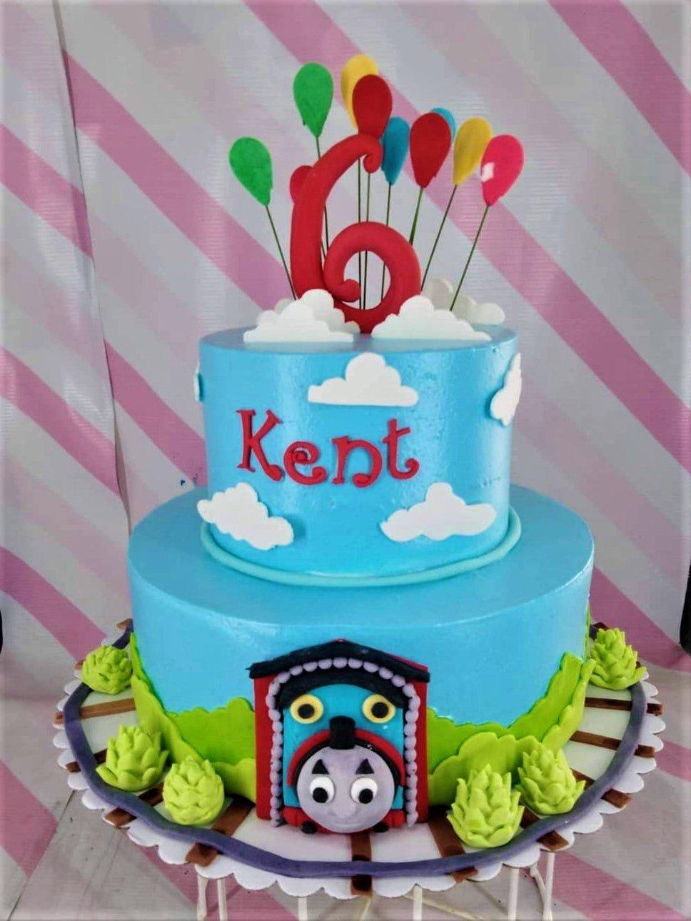 Two-tier blue Thomas the Tank Engine themed birthday cake with a red number 6 topper, colorful balloons, and fondant clouds.