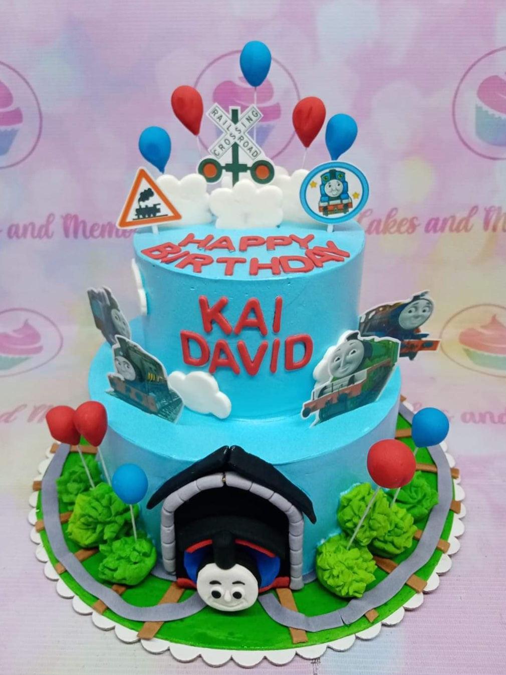 Two-tier light blue Thomas the Tank Engine themed birthday cake with train toppers, balloons, and a tunnel design.