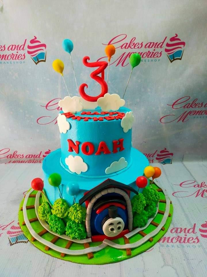Two-tier blue Thomas the Tank Engine themed birthday cake with a 3D train tunnel, fondant clouds, and colorful balloon toppers.