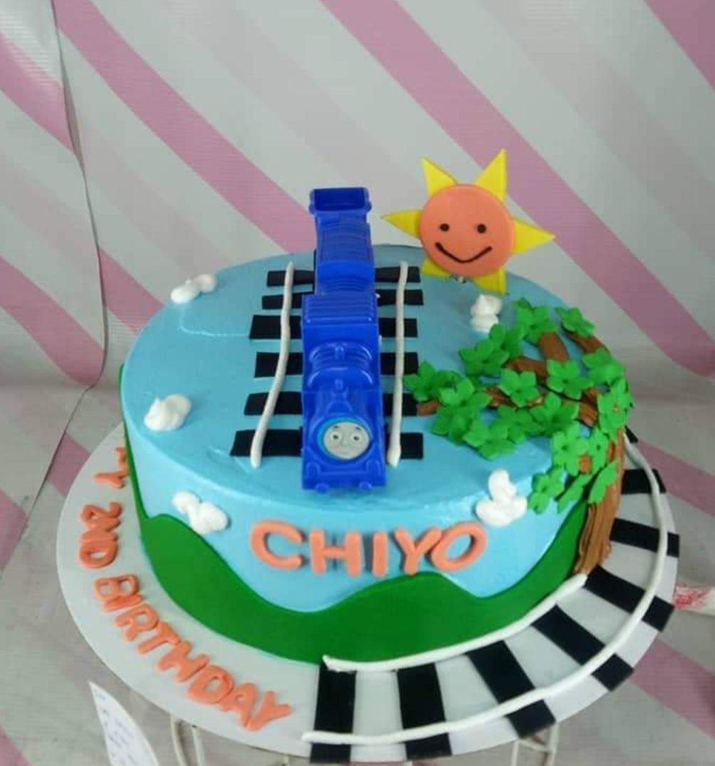 Blue Thomas the Train themed birthday cake with a 3D train topper, fondant sun, and green hills on a track-patterned base.