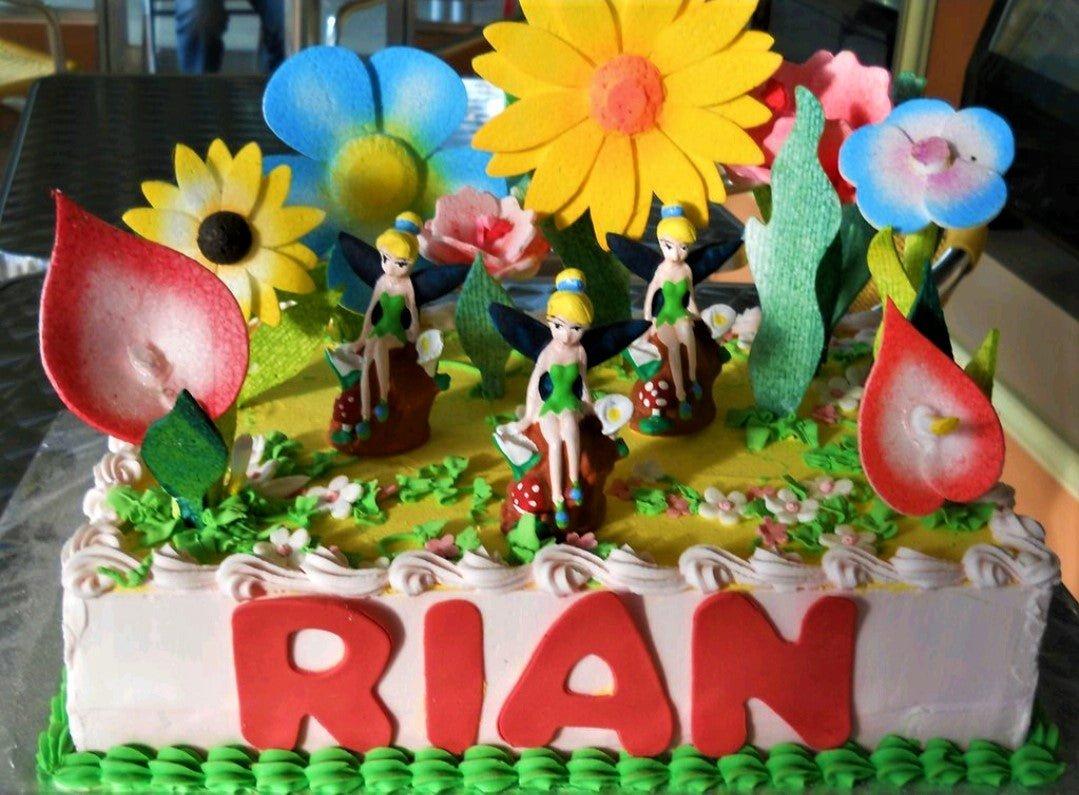 Rectangle Tinkerbell fairy garden cake with large yellow and blue flowers, three fairy figurines, and red gumpaste letters spelling RIAN.