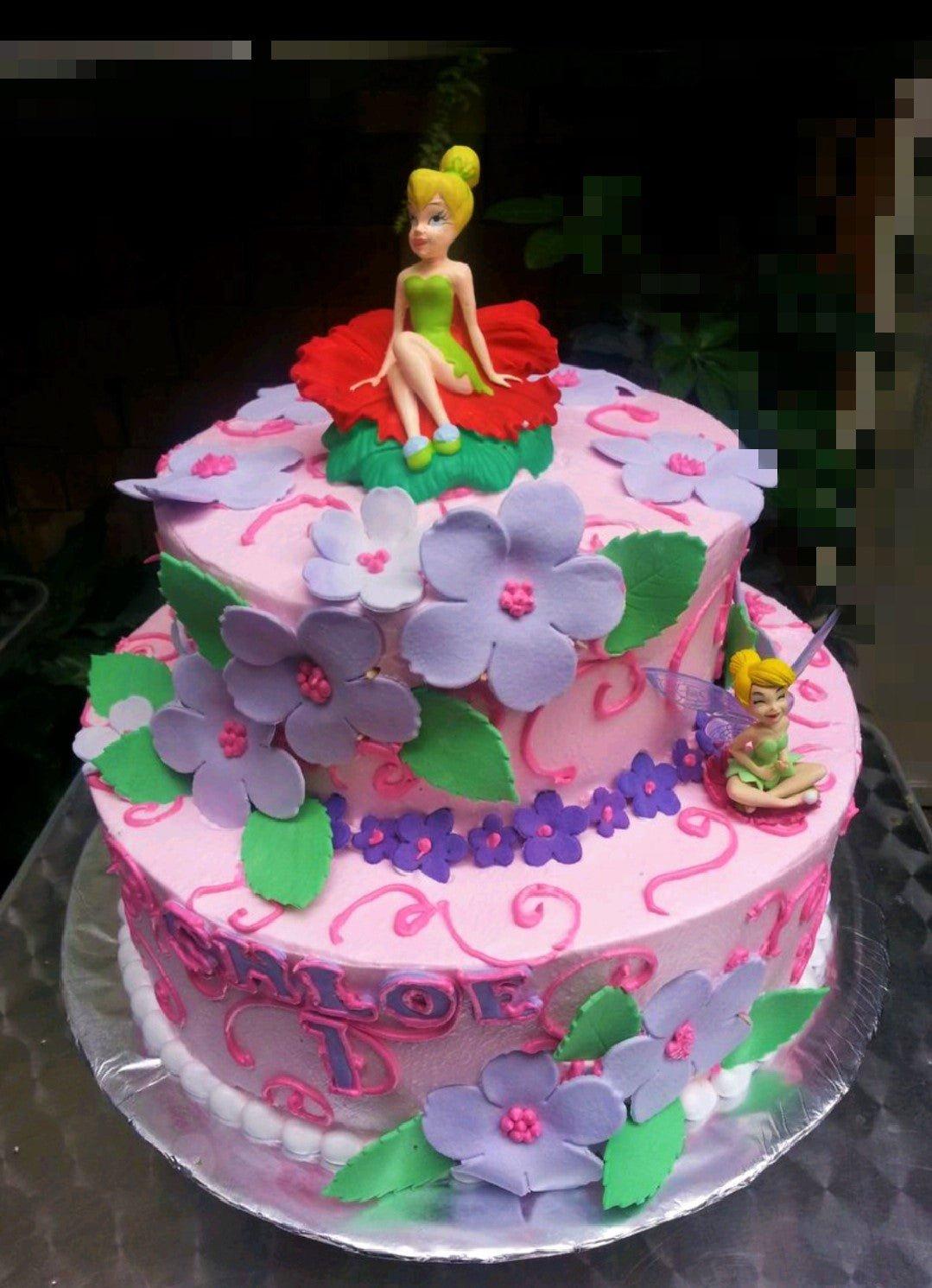 Two-tier pink Tinkerbell themed cake with purple fondant flowers, green leaves, and two Tinkerbell figurines.