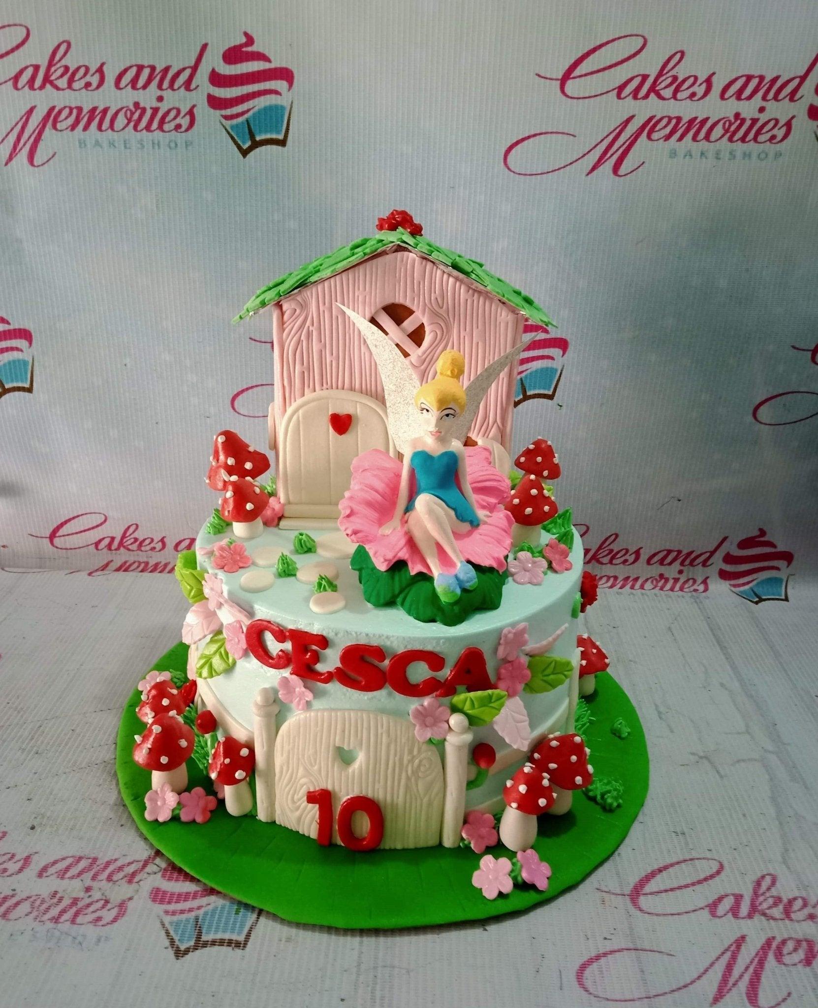 Tinkerbell fairy garden cake with a pink fondant house, red mushrooms, and a Tinkerbell figure sitting on a pink flower.