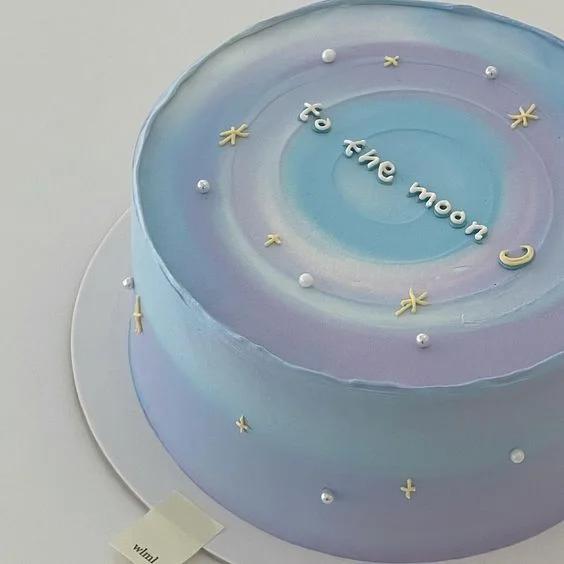 Minimalist 1-tier cake with blue and purple gradient icing, silver dragees, and yellow icing stars featuring the message 'to the moon'.