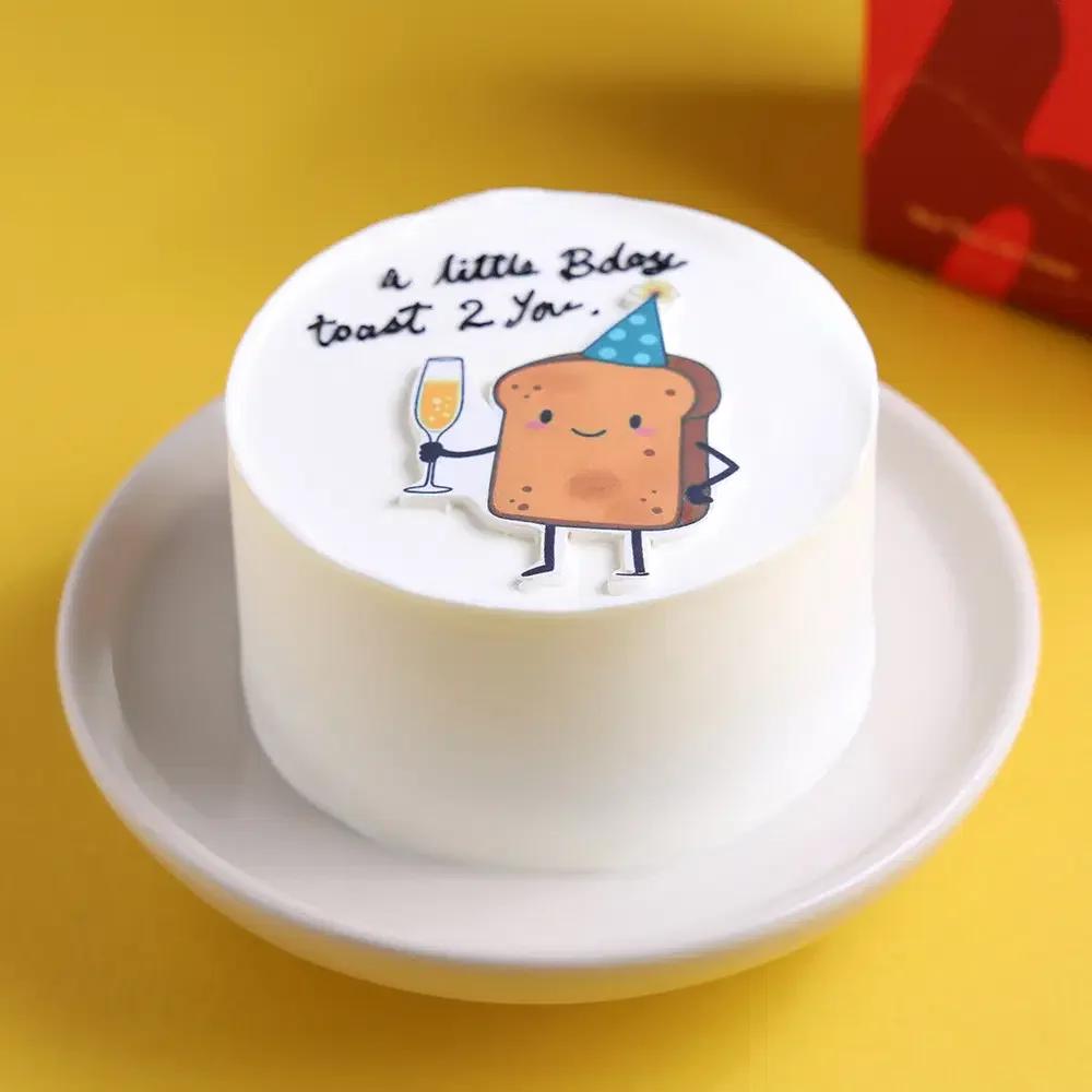 White bento cake with a cute toast character printout holding a champagne glass and a birthday message.