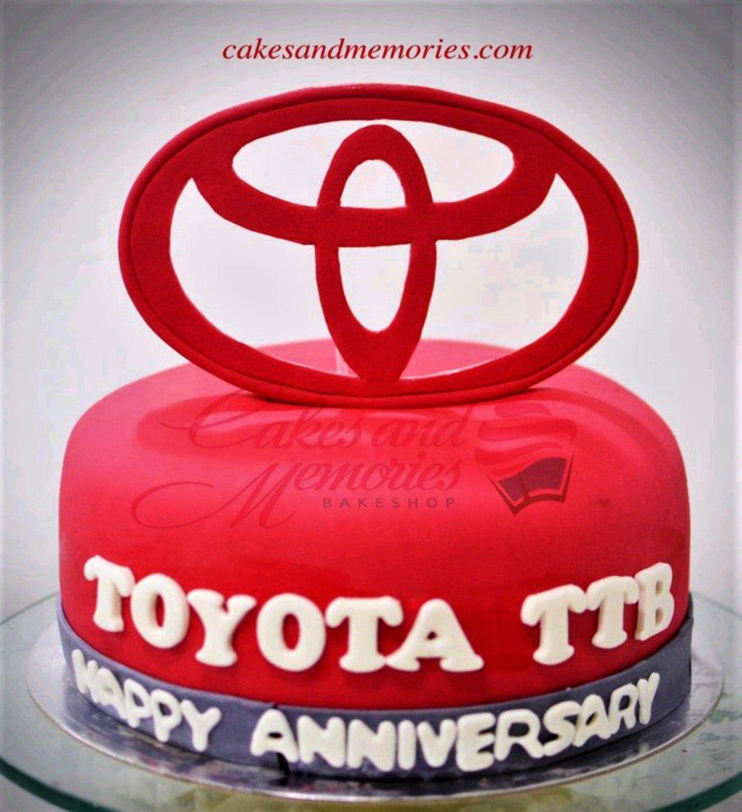 Red fondant cake featuring a large red Toyota logo topper and white gumpaste letters for an anniversary celebration.