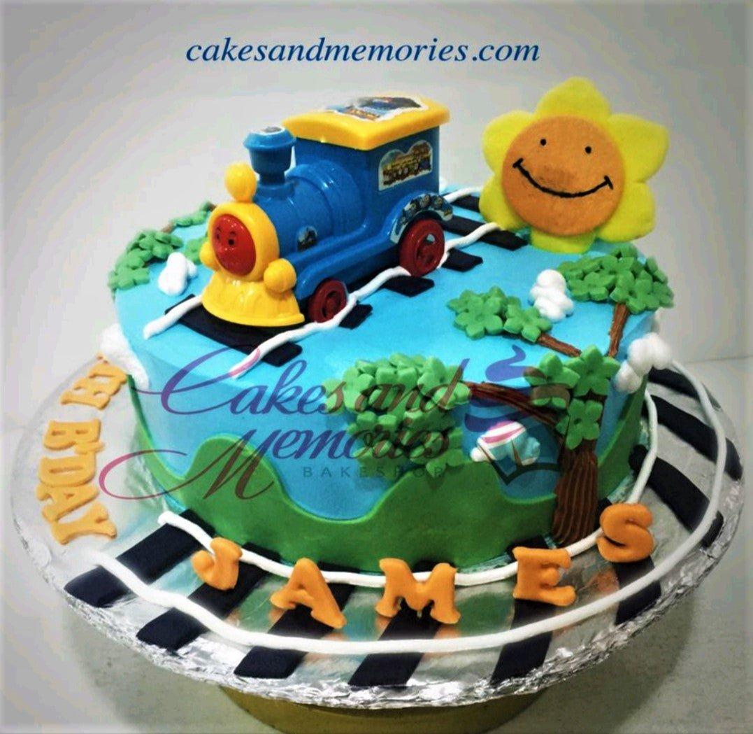 Blue 1-tier train themed birthday cake with a toy train, fondant sun, trees, and a railway track on the base board.