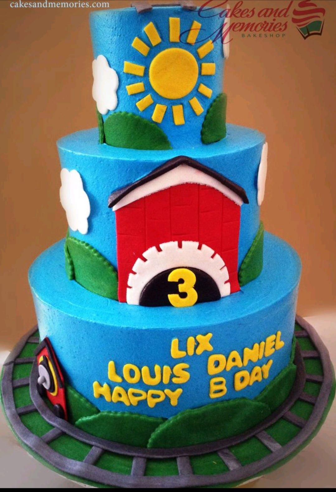 Three-tier blue train-themed birthday cake with a red barn, yellow sun, and train tracks on a green base.