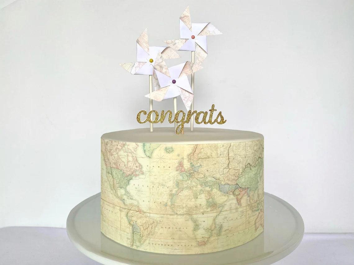 A minimalist white fondant cake featuring a world map side wrap and three map-patterned pinwheel toppers with a gold glitter congrats message.