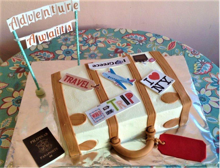 Suitcase-shaped travel themed cake with edible passport, airplane, and travel stickers on a white icing base with brown fondant straps.