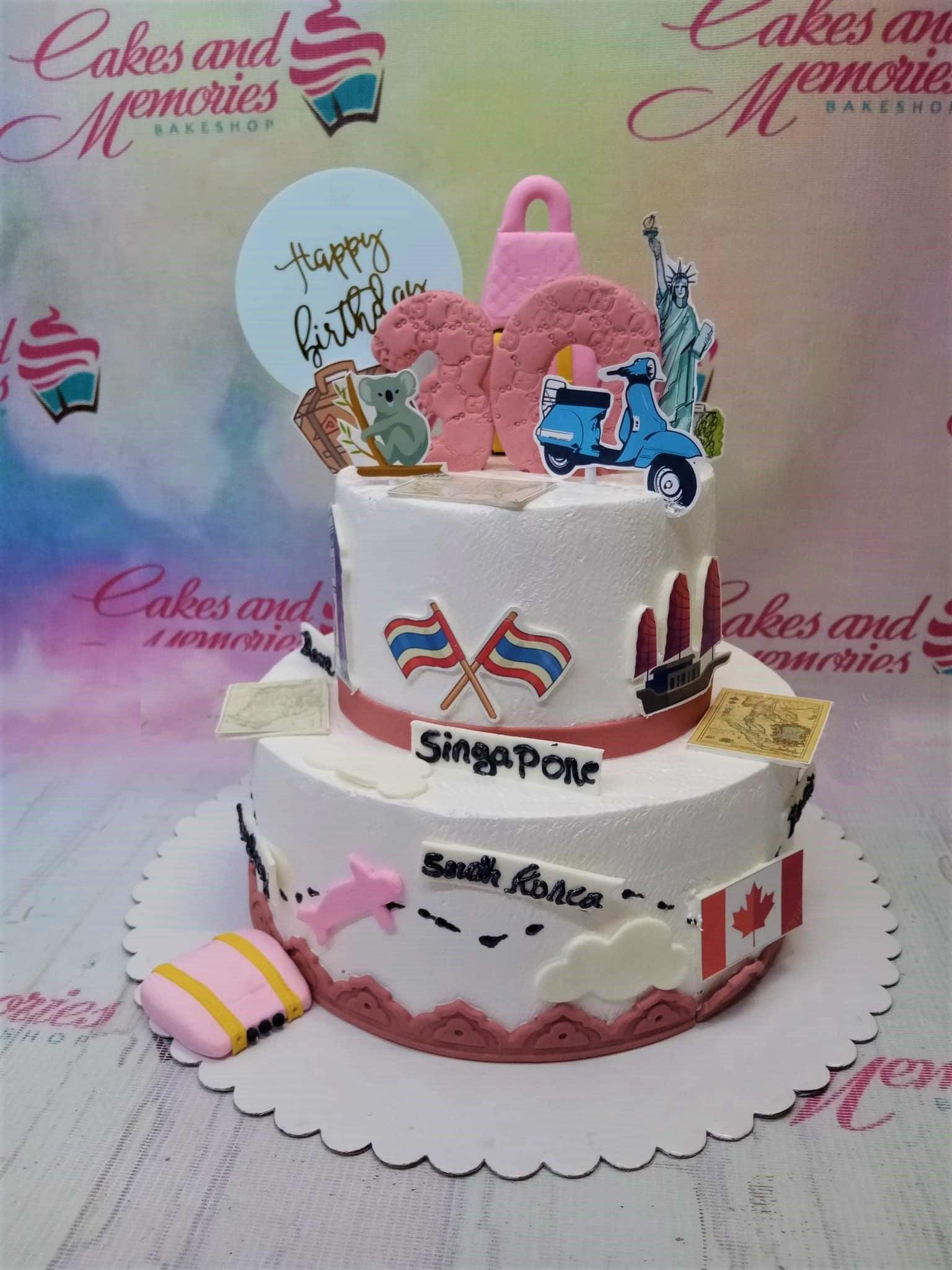 White 2-tier travel themed birthday cake with pink accents, world flags, and travel icons like a scooter and suitcase.