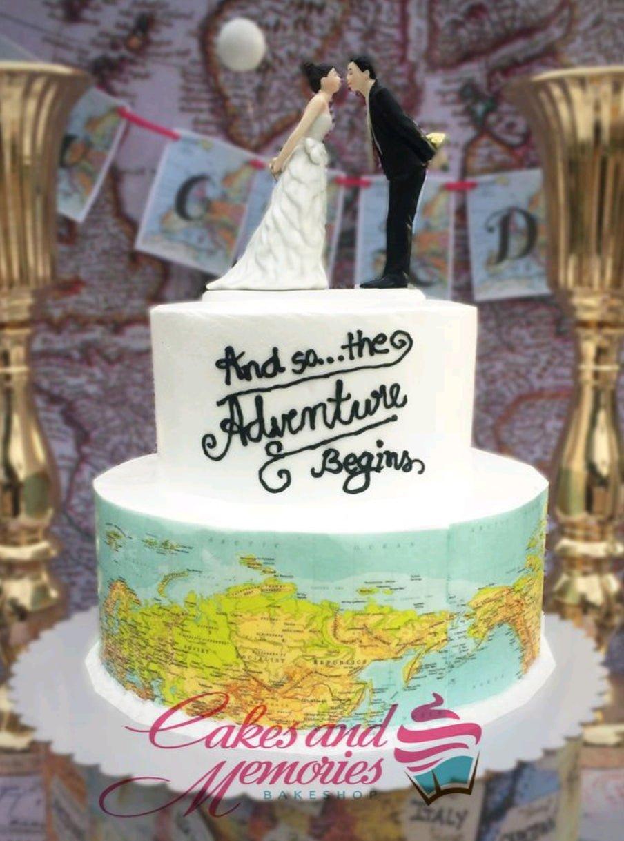 Two-tier white wedding cake with a world map wrap on the bottom tier and a bride and groom topper on top.