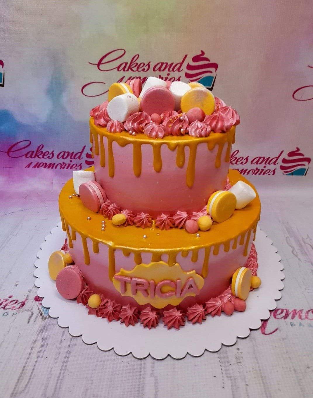 Two-tier pink cake with gold drips, pink and yellow macarons, marshmallows, and a name plaque for Tricia.