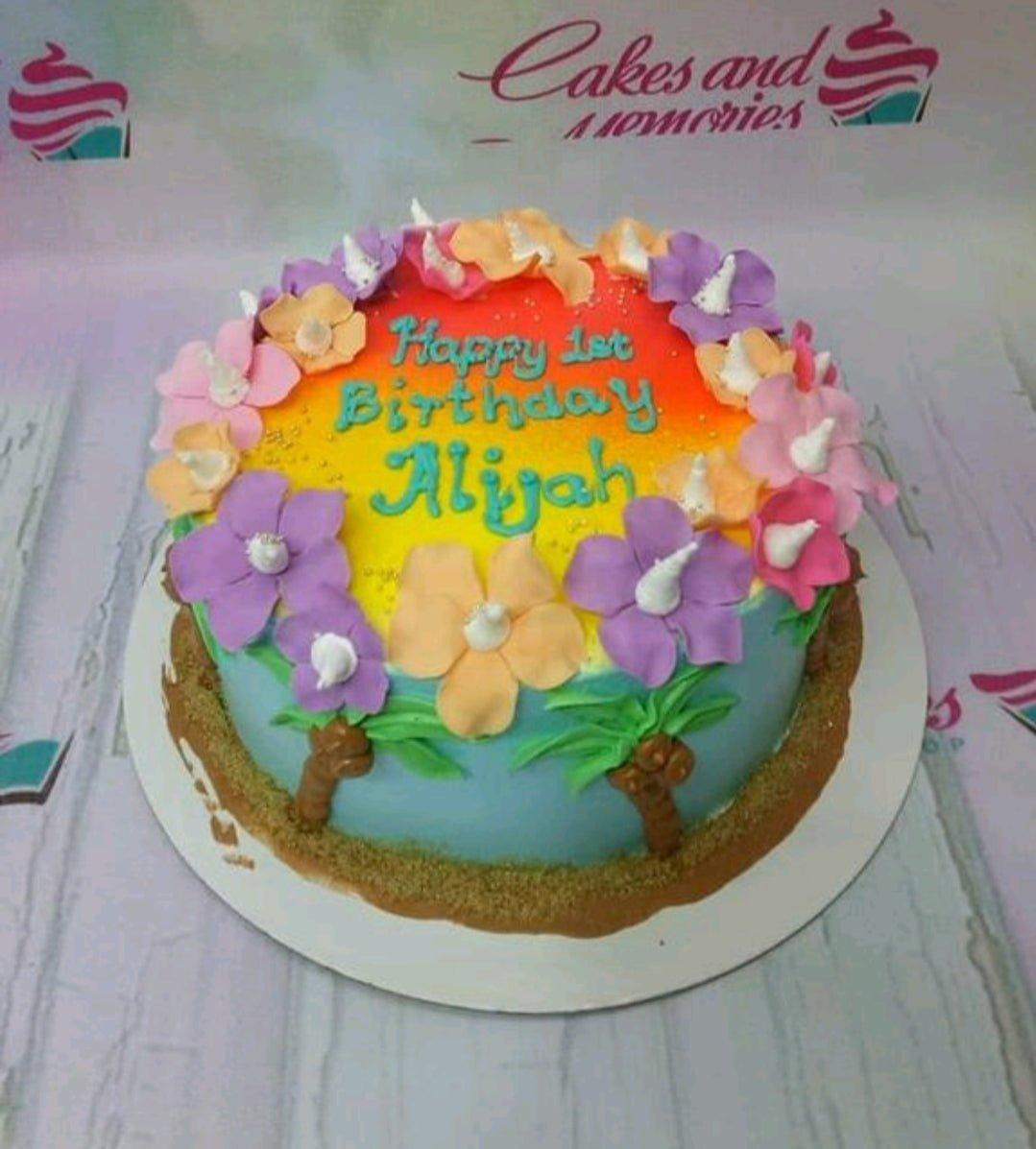Tropical beach themed birthday cake with rainbow sunset icing, hibiscus flowers, palm trees, and a sand-textured base.