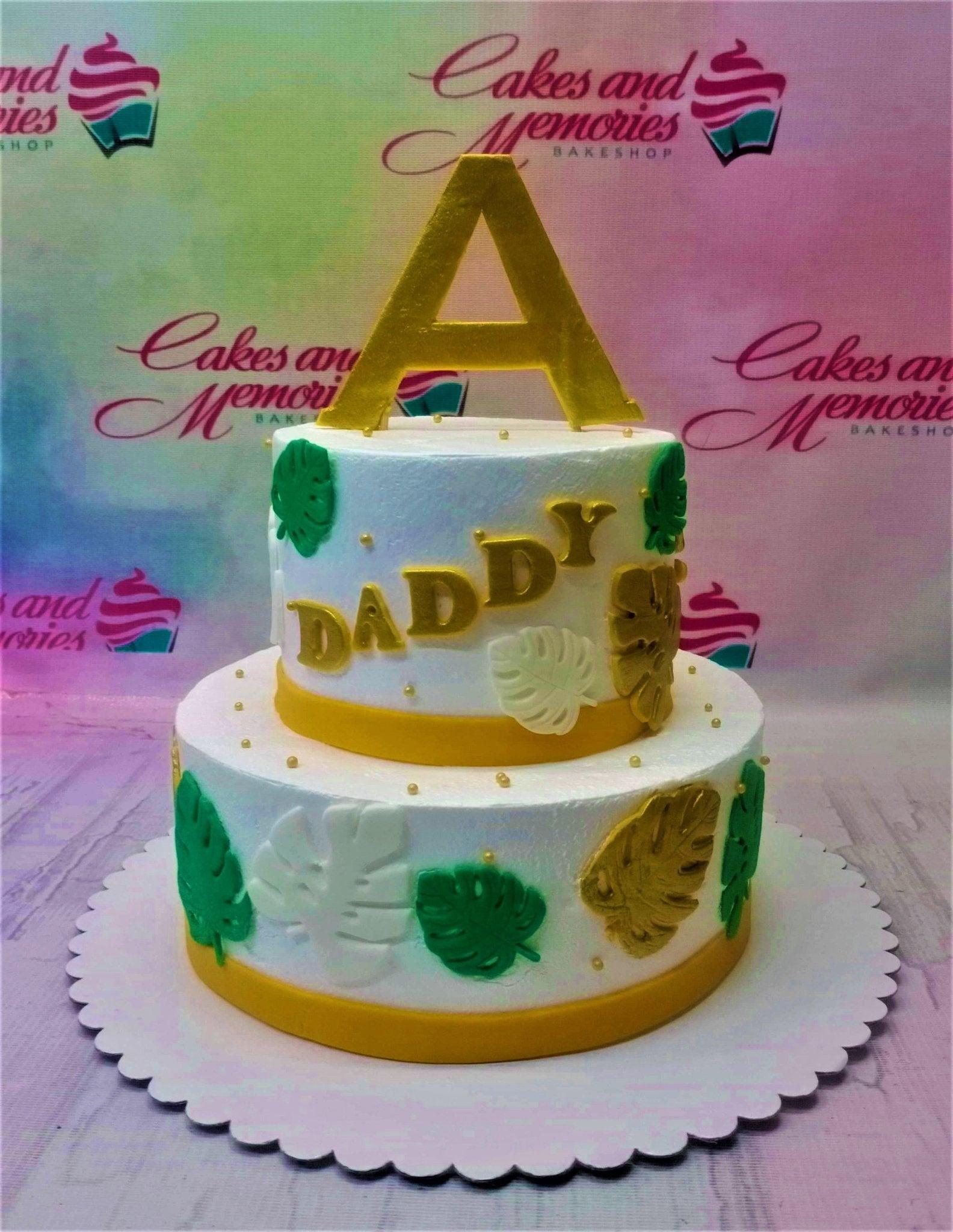 Two-tier white tropical themed cake with gold and green monstera leaves and a large gold letter A topper.
