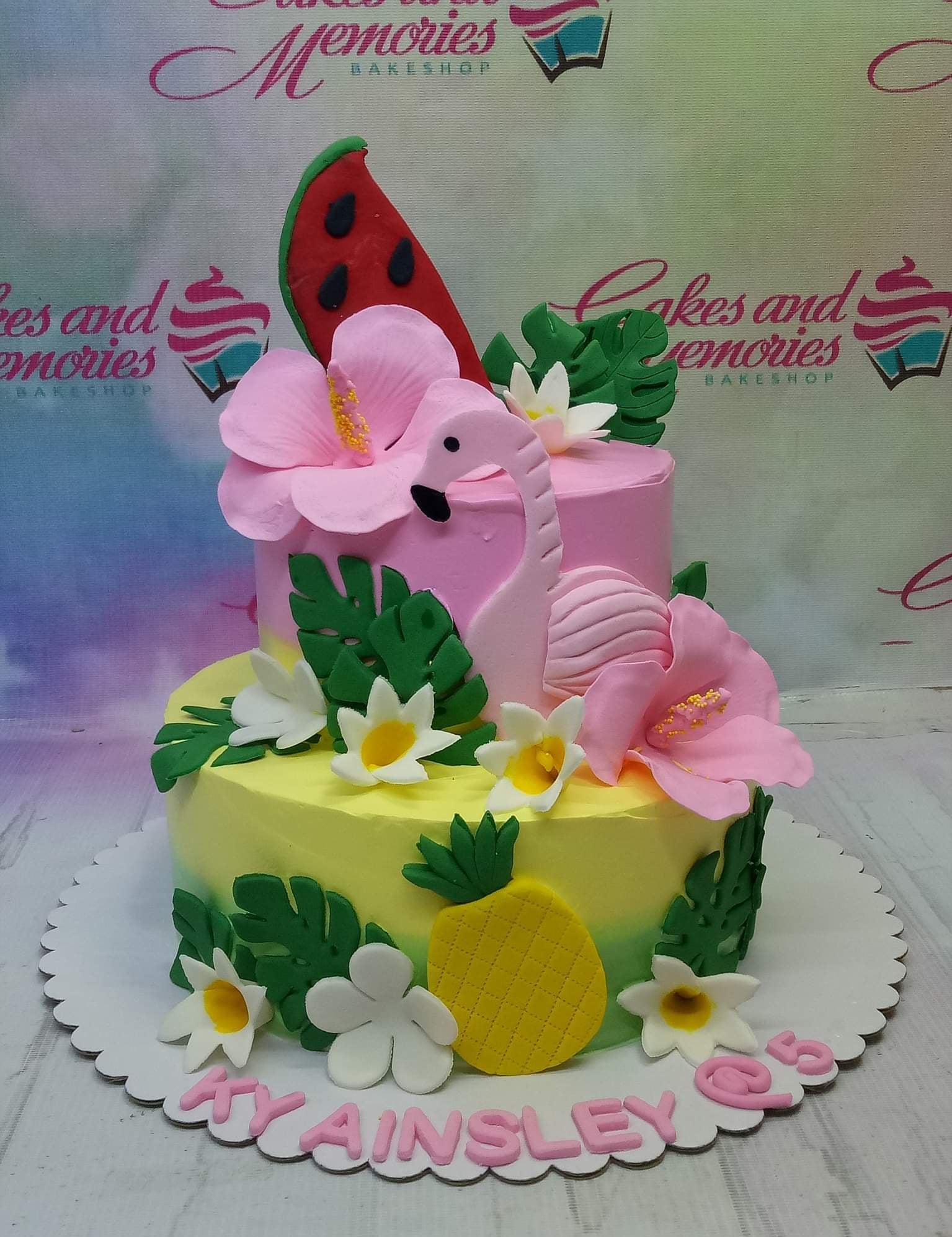 Two-tier tropical flamingo cake with pink and yellow icing, fondant hibiscus flowers, monstera leaves, watermelon, and pineapple toppers.