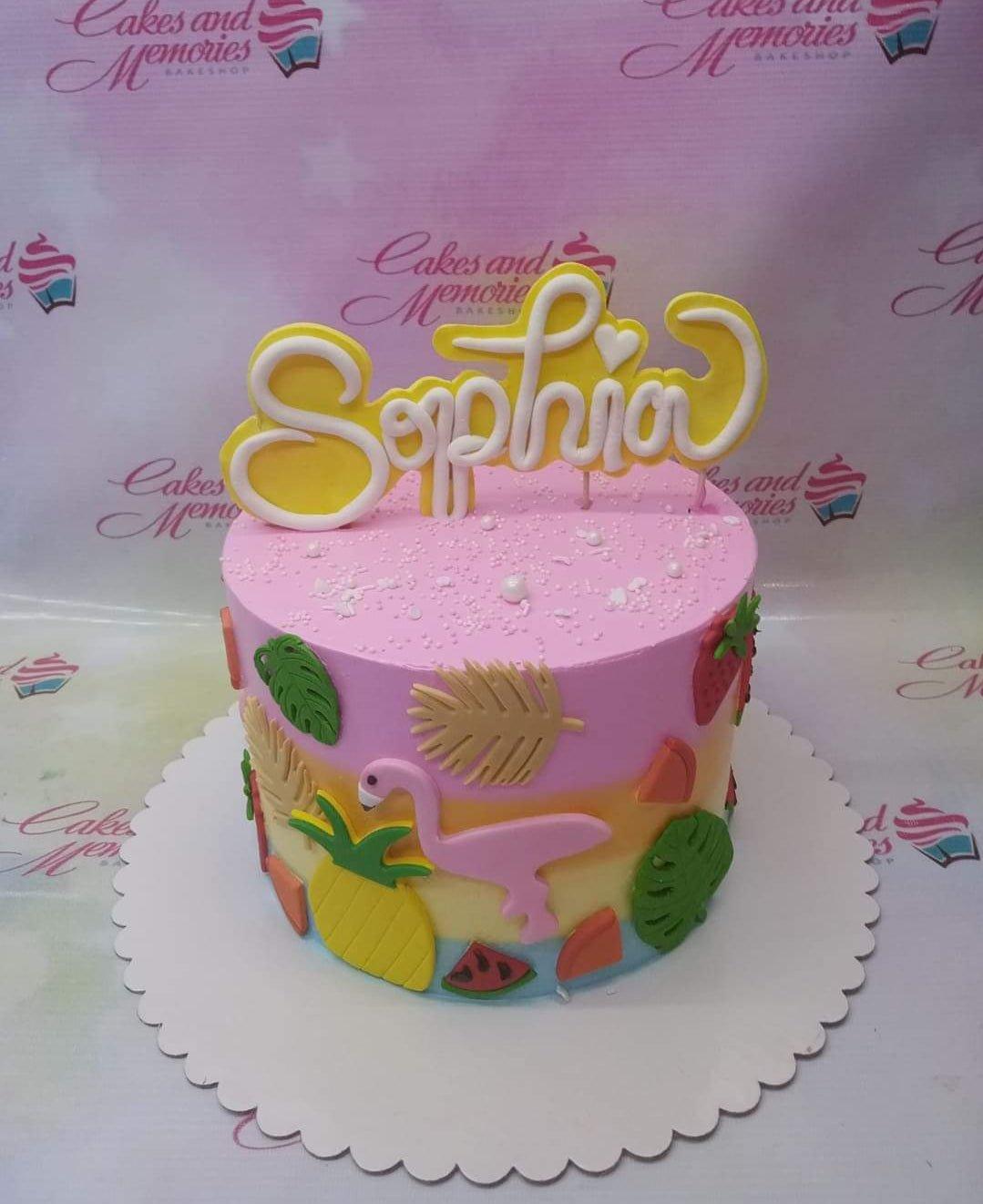 Tropical flamingo and fruit themed pink cake with a large yellow Sophia name topper and fondant tropical leaves and fruits.