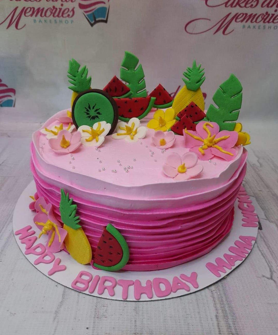 Pink tropical fruit themed birthday cake with fondant watermelon, pineapple, kiwi, and hibiscus flowers.