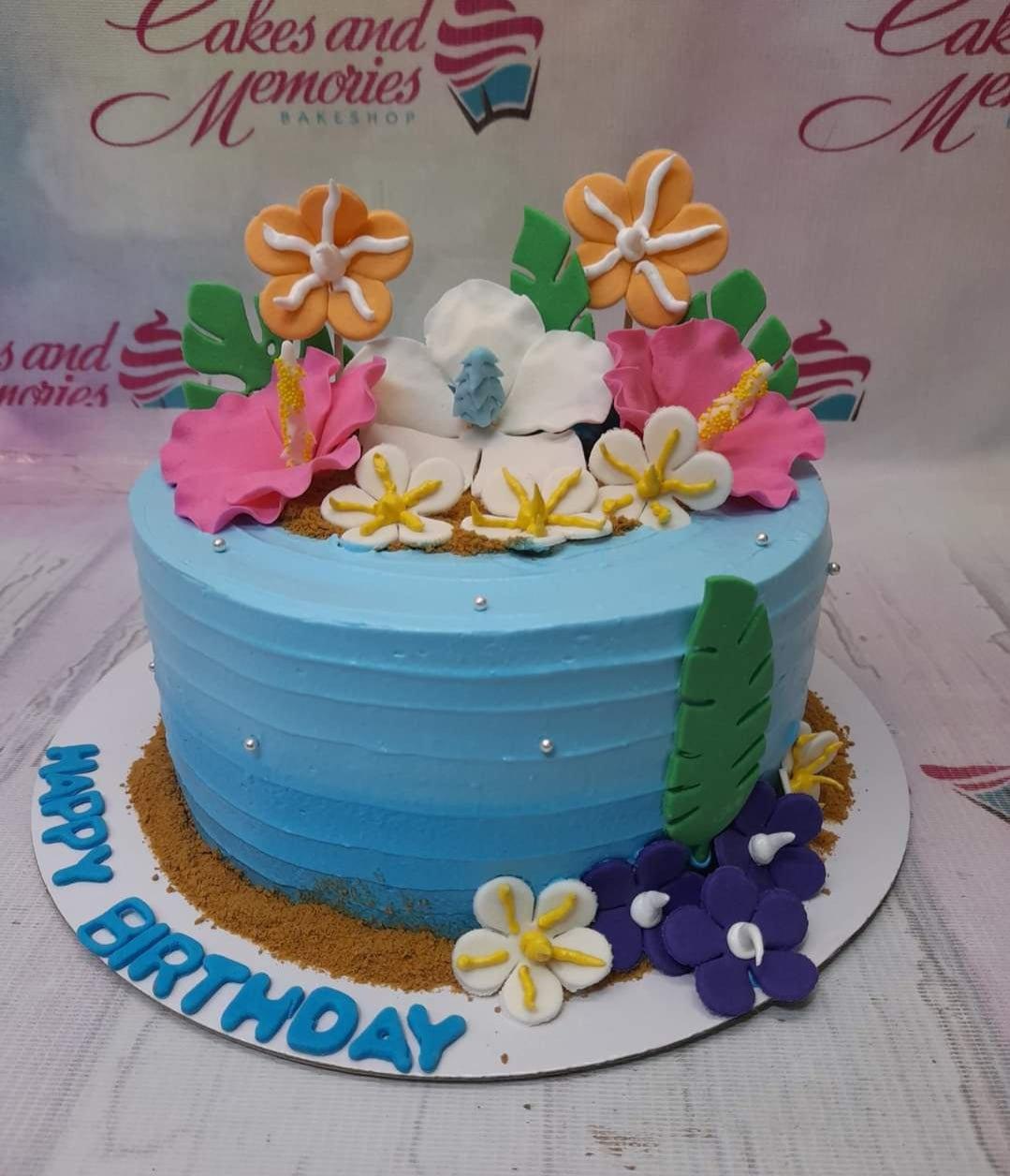Tropical blue cake with edible hibiscus flowers, green leaves, and brown sugar sand base for a beach-themed birthday celebration.