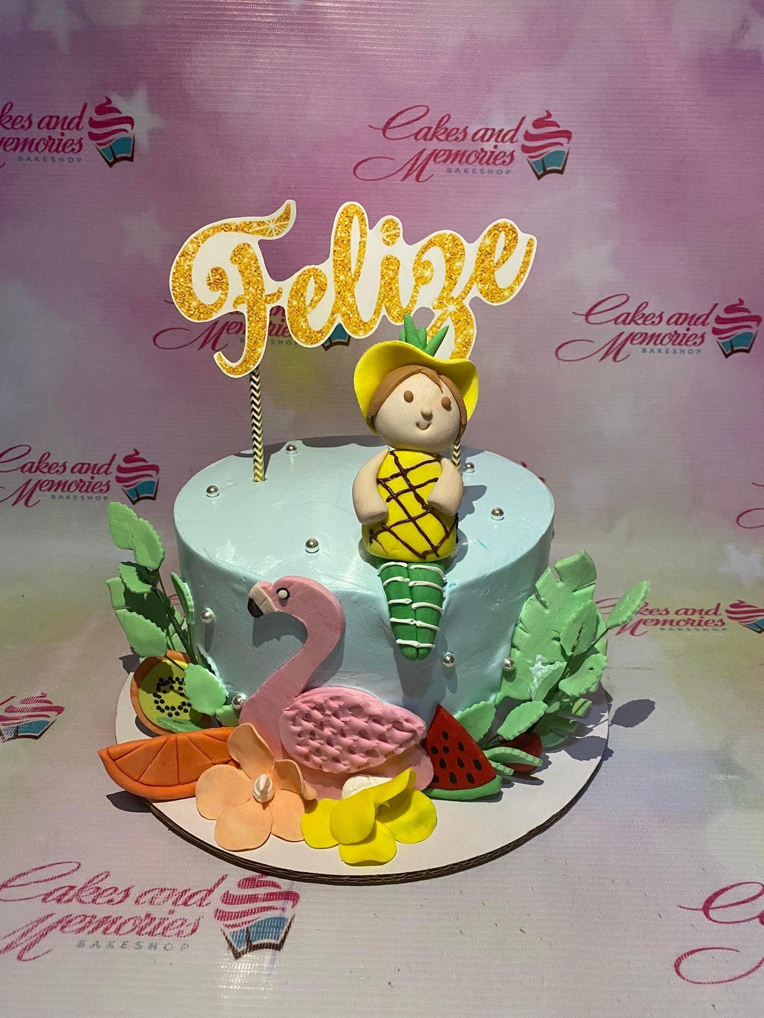 Light blue tropical themed cake with a fondant girl in a pineapple outfit, a pink flamingo, and various fruit slices.