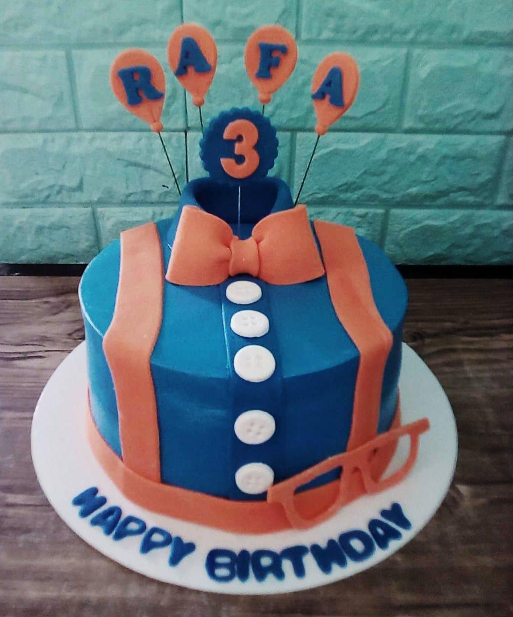 Blue tuxedo-themed birthday cake with orange suspenders, a bow tie, and balloon-shaped toppers for a 3rd birthday.