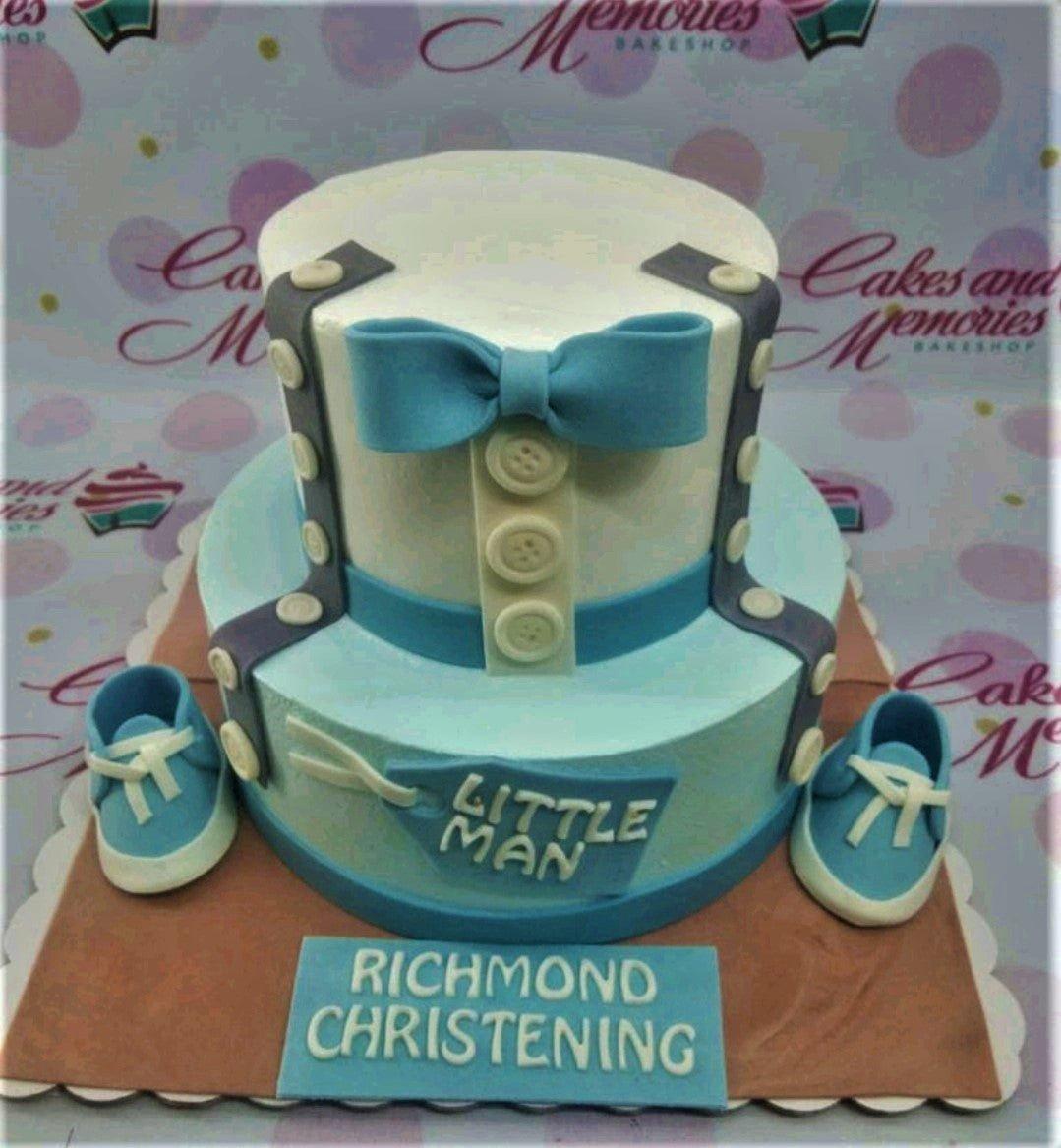Two-tier light blue and white tuxedo-themed christening cake with a blue bow, buttons, and fondant baby shoes.