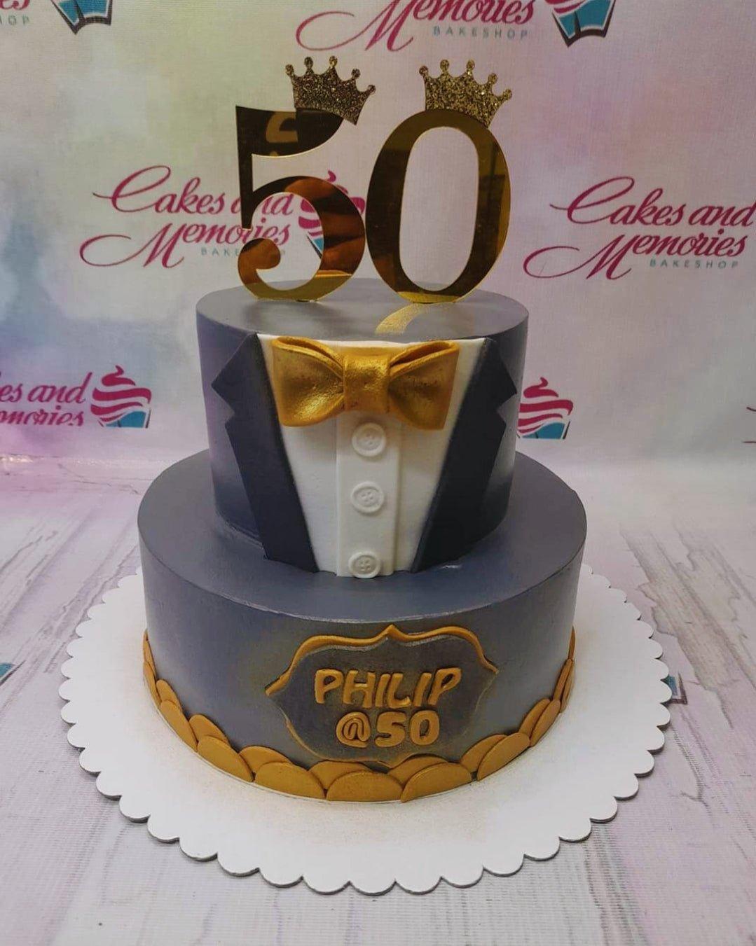 Two-tier gray tuxedo-themed 50th birthday cake with gold bow tie and large gold number 50 toppers.