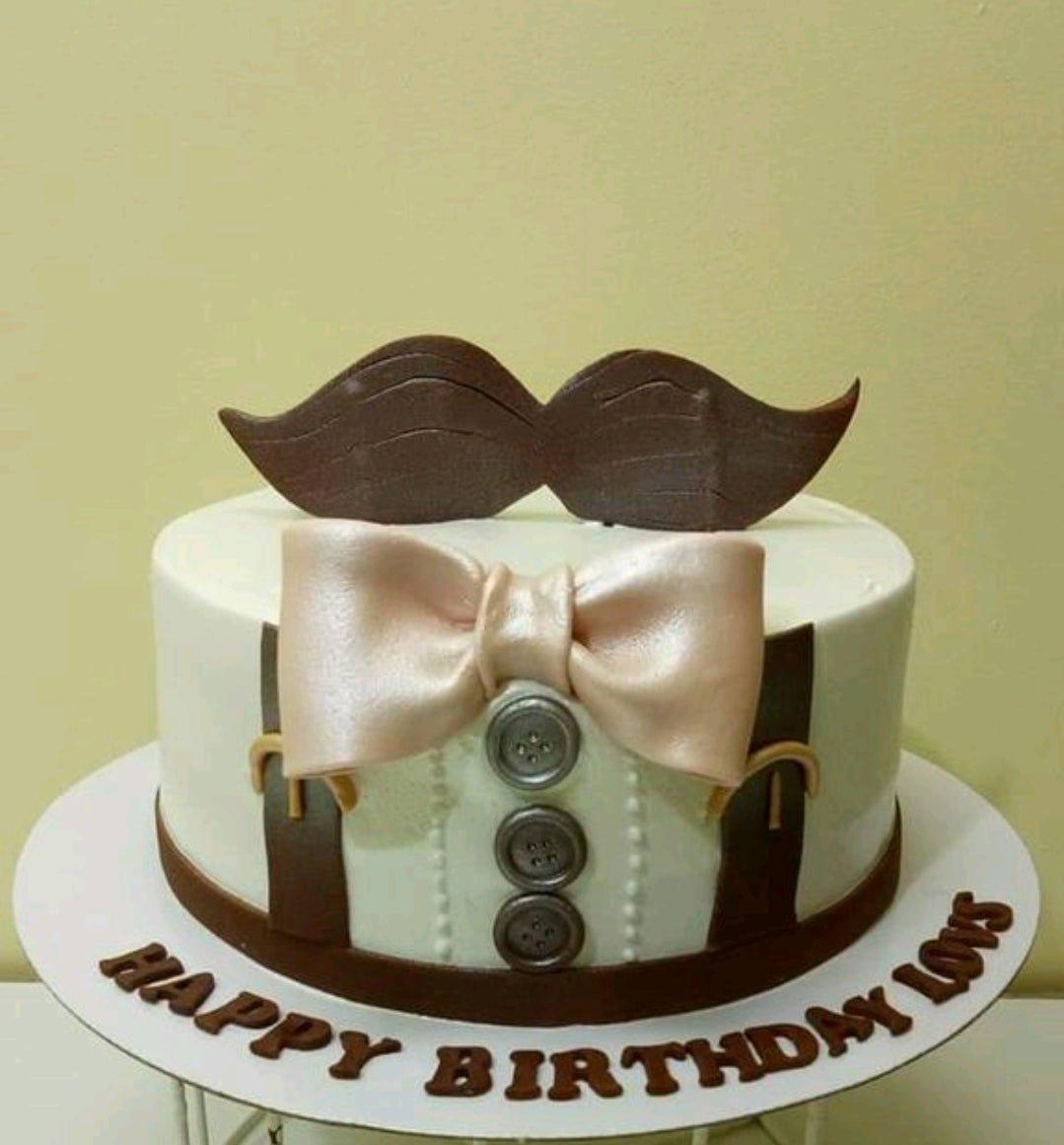 White fondant tuxedo cake with a large brown mustache topper, a rose gold bow tie, and brown suspenders with buttons.