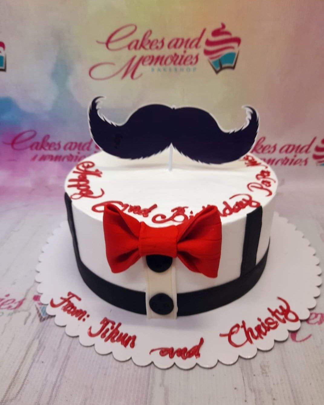 White fondant tuxedo cake with a red bow tie, black buttons, and a large black mustache topper for a 2nd birthday celebration.