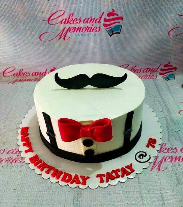 White tuxedo-themed birthday cake with a black mustache topper, red bow tie, and black suspenders on a white scalloped board.