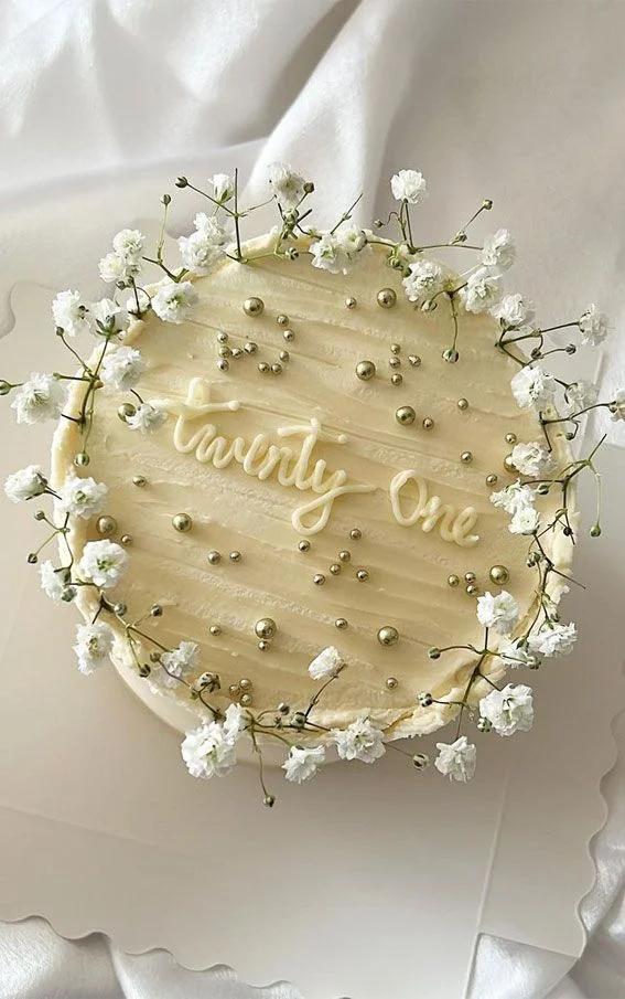 Elegant white minimalist cake with fresh baby's breath flowers, gold dragees, and 'Twenty One' piped icing message.