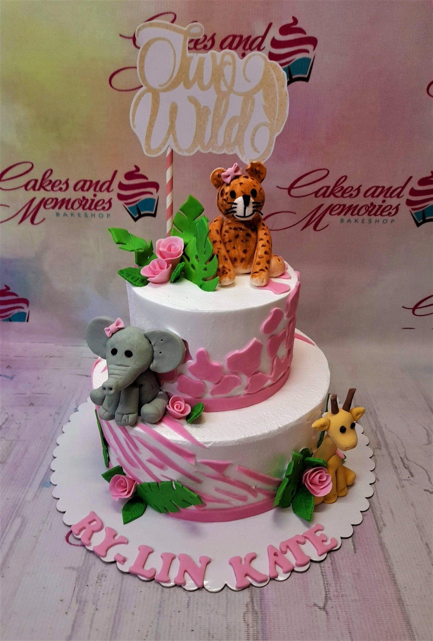 Two-tier white jungle safari cake with pink animal print, fondant leopard, elephant, and giraffe figures, and a 'Two Wild' topper.