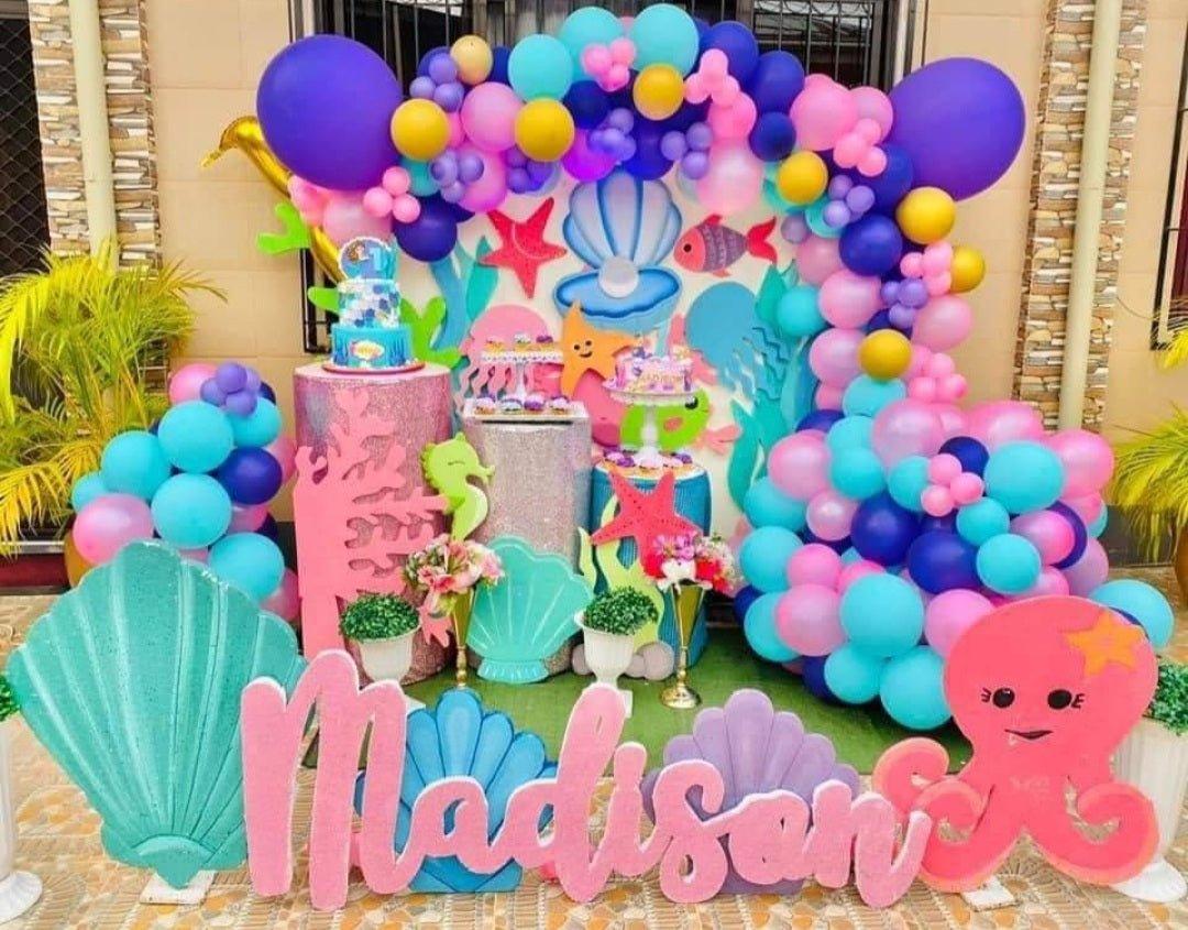 Two-tier under the sea themed birthday cake with blue icing, mermaid tail, and sea creature toppers on a colorful balloon backdrop.