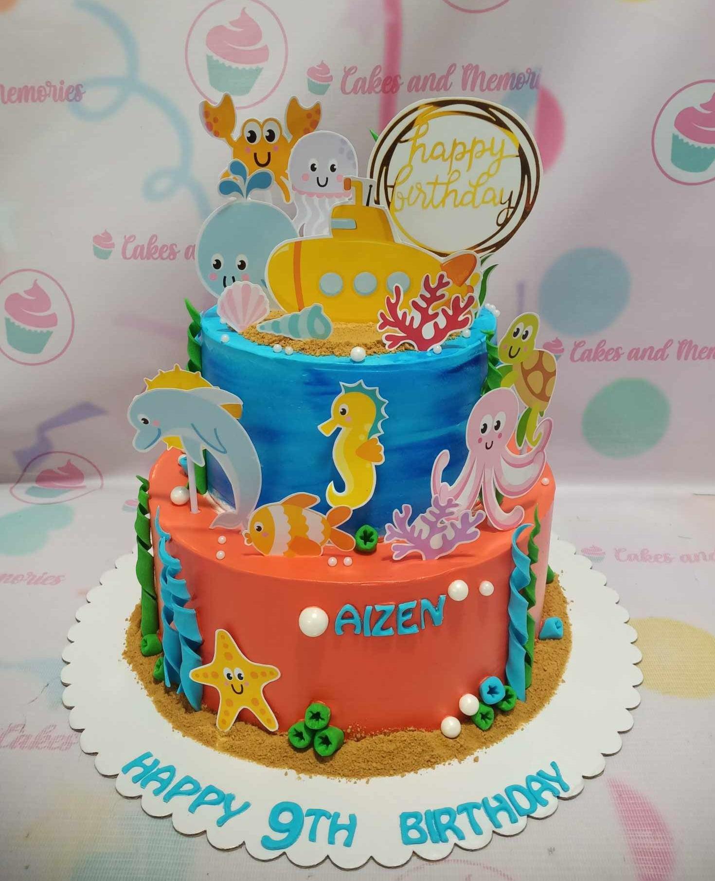 Two-tier under the sea themed birthday cake with blue and orange icing, featuring various sea creature printouts and a yellow submarine topper.