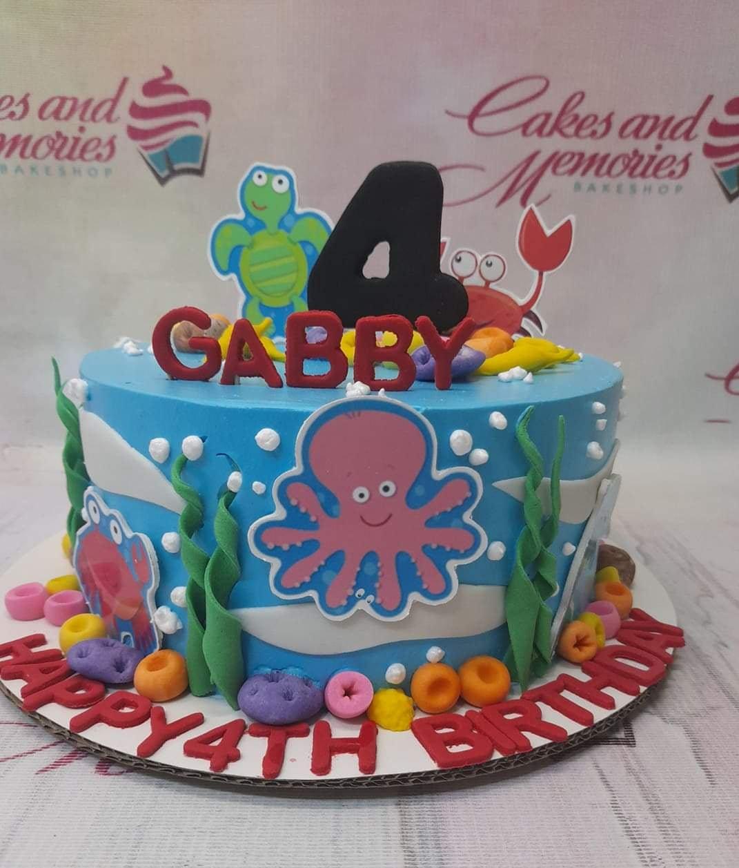 Blue under the sea themed birthday cake with a pink octopus, turtle, and crab printouts, featuring red gumpaste letters for Gabby's 4th birthday.