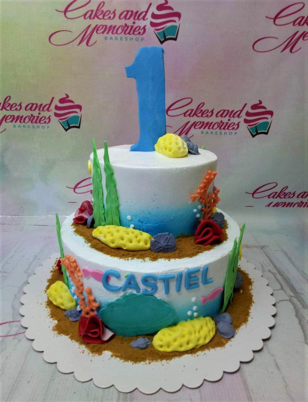 Two-tier under the sea themed birthday cake with blue gradient icing, fondant coral, seaweed, and a large blue number one topper.