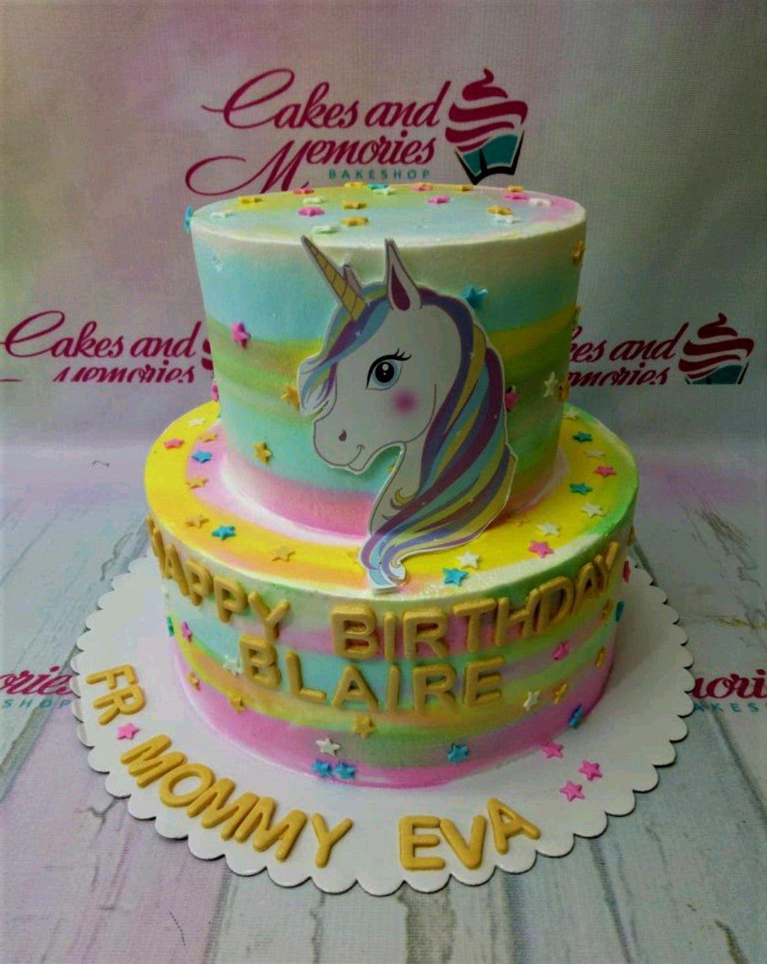Two-tier rainbow unicorn birthday cake with a large unicorn printout, gold gumpaste letters, and colorful star decorations.