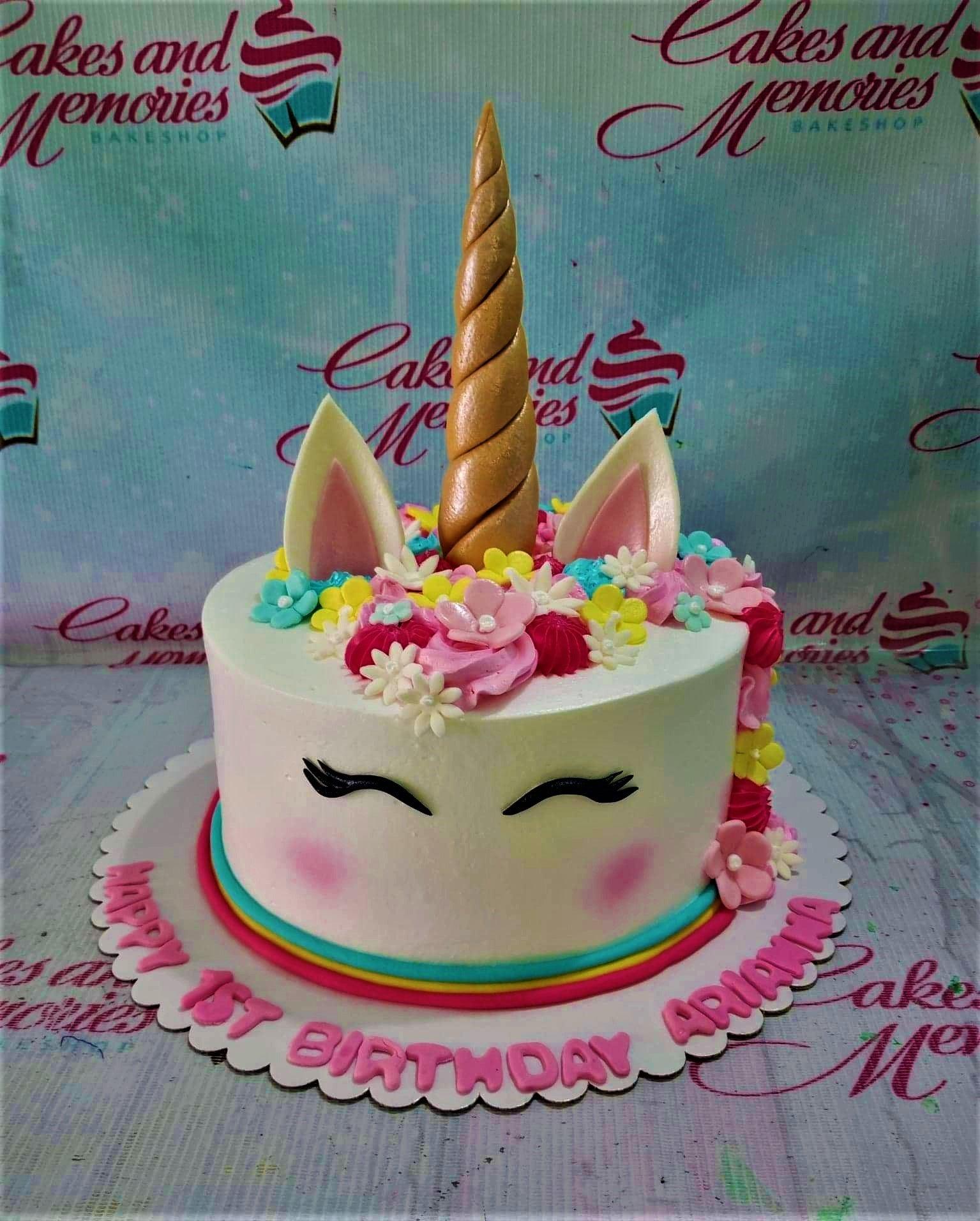 White unicorn cake with a gold horn, pink ears, and a colorful floral mane of pink, yellow, and teal icing flowers.