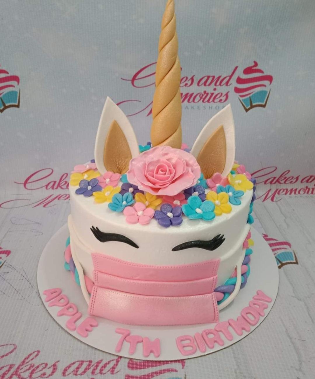White unicorn birthday cake with a pink face mask, gold horn, and colorful fondant flowers.